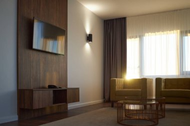 Duvara monte edilmiş televizyon, ahşap mobilyalar ve akmakta olan perdelerle dolu geniş pencereli modern oturma odası sıcak bir ortam ve davetkar bir atmosfer yaratıyor.