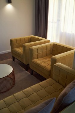 İki sarı koltuk ve yumuşak tavan lambasının altında yuvarlak bir sehpanın olduğu rahat bir oturma odası. Pencere perdelerinden süzülen doğal gün ışığı sıcaklık katıyor.