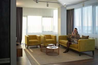 Modern ofiste koltukta oturan iş kadını kucağında dizüstü bilgisayarla telefonda konuşuyor ve önünde kahve masası var. Arkaplanda büyük pencereler ve sarı koltuklar