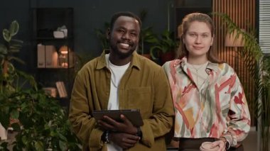 Modern ofis alanında elinde dijital tablet ve kahve bardağıyla kameraya poz veren iki çift ırklı Z işçisinin orta boy portresi.