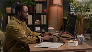 Z jenerasyonunun orta boy portresi Afrikalı Amerikalı erkek programcı dijital cihazlarda kodlama yapıyor ve eko ofis alanındaki kameraya bakıyor