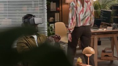 Sanal gerçeklik gözlüğü kullanan genç Afrikalı Amerikalı adama odaklanırken, beyaz kadın meslektaşı da modern bilişim ofisindeki bilgisayar ve kodlama ekranlarıyla masada oturuyor.