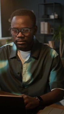 Laptopun önünde oturan ve ofis masasındaki kameraya gülümseyen Afrikalı Amerikalı erkek çalışanın dikey portresi.