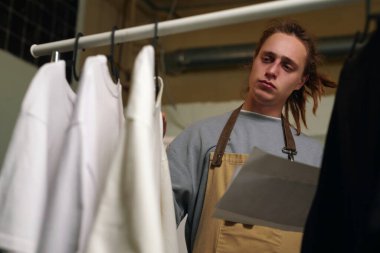 Düşük açılı, elinde kağıtlar olan ve askıdaki büyük boy tişörtlere bakan yetişkin bir adamın fotoğrafı.