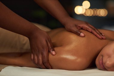 Bir terapistin elleri masaj masasına uzanmış hastasının omuzlarına dikkatlice masaj yapıyor, mum ışığı ve bulanık arka plan huzur ve huzur verici bir ortam yaratıyor.