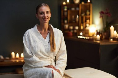 Beyaz cüppeli sakin bir kadının portresi. Etrafı mumlar ve yumuşak aydınlatmalarla çevrili sakin bir spa ortamında oturuyor.