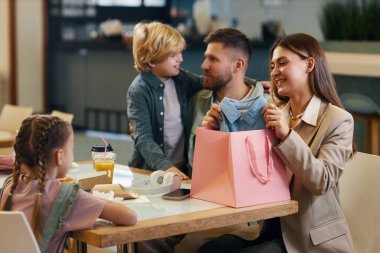 Güzel, genç bir anne, küçük kızıyla kız gibi sohbet ederken kocaları da bulanık arka planda sarışın oğluna sarılıyor.