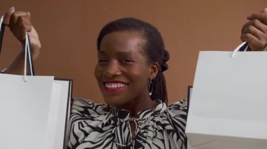 Kahverengi arka planda alışveriş torbalarıyla poz verirken gülümseyen mutlu siyahi kadının orta boy portresi.