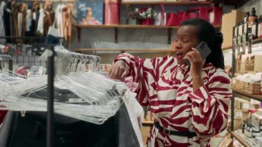 Moda mağazasında giysi seçerken cep telefonuyla konuşan neşeli siyahi kadın müşterinin orta boy fotoğrafı.