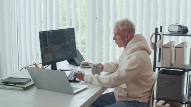 Bilgisayarlar ve dizüstü bilgisayarlar için uygulama geliştirirken çay içen genç bir erkek yazılımcının yan görüntüsü, ofis masasında oturuyor.