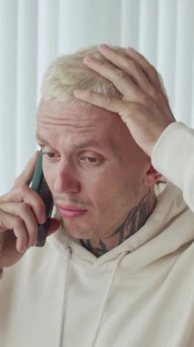 Boynunda dövme olan sinirli bir beyaz adamın evde telefonla konuşurken dikey çekimi.