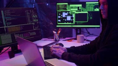 Siyah kapüşonlu genç beyaz adam karanlık odada şifreli senaryolarla bilgisayarları hackliyor.