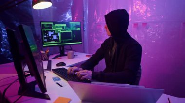 Erkek siber suçlunun karanlık odada bilgisayarlardaki kodlama ekranlarıyla otururken kredi kartlarından veri çalarken orta ölçekli görüntüsü.