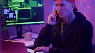 Boynunda dövmesi olan beyaz bir adamın laptopta programlama yaparken cep telefonuyla konuşurken, sunucu neon odasını hacklerken görüntüsünü kaldır.