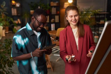 Bir kadın, tablet bilgisayar kullanırken meslektaşı ile birlikte çizelge üzerinde yazıyor ve proje hazırlıyor.
