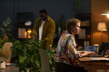 Bilgisayarda güvenlik kodlarıyla çalışan genç bir kadın ve arka plandaki yeşil bitkileri önemseyen bir adam.