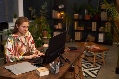 Genç iş kadını, modern ofiste yeşil bitkilerle masada otururken bilgisayarla çalışıyor.