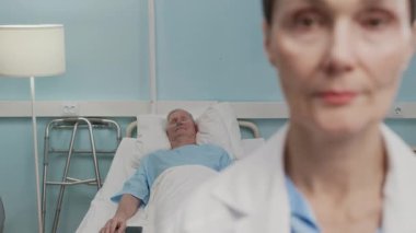 Yetişkin beyaz kadın pratisyen hekimin portresi hastane koğuşunda yatakta yatan yaşlı erkek hasta kameraya bakarken kameraya bakıyor.