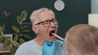 Klinikteki yaşlı bir hastanın boğazını incelerken spatula kullanan erkek pratisyen hekimin el kamerası görüntüsü.