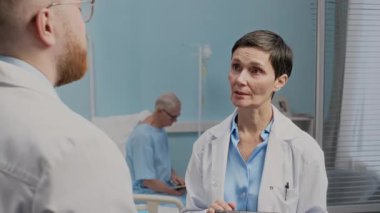 Hastane odasında yaşlı hastaların tedavisini tartışırken beyaz önlüklü iki beyaz doktorun dijital tablet kullanırken orta boy fotoğrafı.