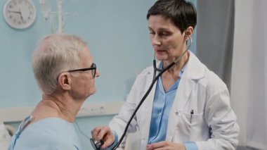 Hastanenin koğuşunda steteskop kullanarak yaşlı hastaları muayene eden orta yaşlı bir kadın.