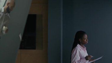 Genç Afrikalı kadın yöneticinin, elinde bir dosya tutarak modern ofis alanındaki beyaz erkek meslektaşıyla konuşmasının el kamerası görüntüsü.