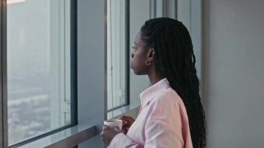 Orta boy pembe tişörtlü, kahve içen, modern binadaki sisli şehir manzarasına bakan siyahi bir kadının fotoğrafı.
