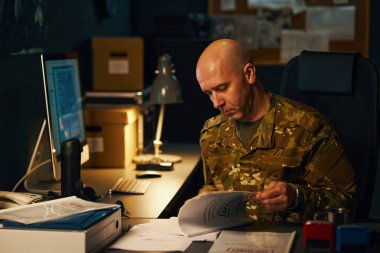 Kamuflaj üniforması giyen ordu subayı bilgisayar ve lambayla ofis masasındaki belgeleri okuyor ve analiz ediyor. Evraklar ve ofis malzemeleri masaya saçılmış.