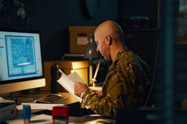 Askeri üniformalı biri bilgisayar ekranlı kağıtlara yoğunlaşıyor.