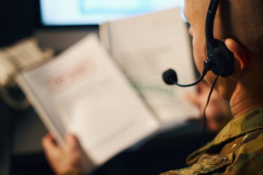 Kamuflaj üniforması giyen, el kitabı okuyan ve odaklanmış bir ortamda özel kulaklık kullanan kişiler.