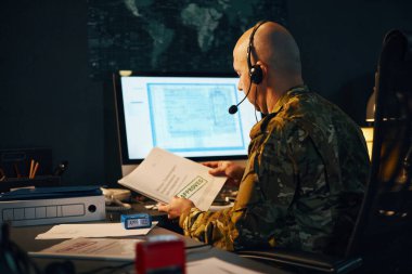 Ofiste çalışırken elinde bir belge tutan askeri kıyafetli bir adam.