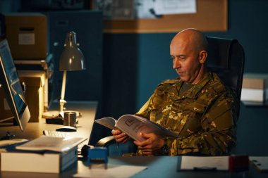 Kamuflaj üniformalı ordu subayı ofis ortamında masa lambası altında evrak okuyor ve arka planda raflar ve bilgisayar var.
