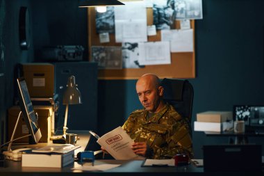 Askeri üniformalı bir adam gece geç saatlerde ofiste çalışıyor, önemli belgeleri inceliyor ve masa lambasıyla aydınlatılmış çalışma alanındaki görevlere odaklanıyor ve göreve hazırlanıyor.