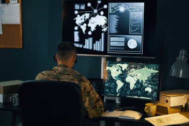 Üniformalı asker, bir komuta merkezindeki çoklu dijital ekranların küresel verilerini ve bilgilerini analiz ediyor. Masada oturan adam ekranlarla aydınlatılmış önemli görevlere odaklanıyor.