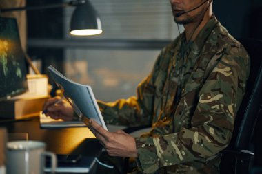 Askeri üniforma giymiş bir kişi, bir ofis masasında, üzerinde bir kulaklık varken, loş ışıklandırılmış çalışma alanında odaklanmış ve özenli bir tavır sergiliyor.