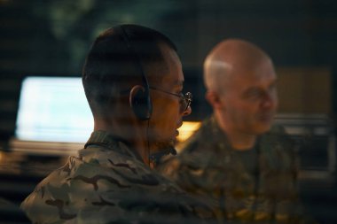 Komuta merkezi içinde gözlem yapan iki ordu subayı. Bir memur kulaklık takıyor ve elindeki işe odaklanmış görünüyor.