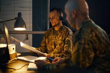 Ordu personeli masa lambalarının altında masalarında çalışırken kulaklık kullanarak hassas belgeleri tartışıyor.