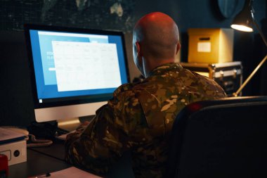 Masaüstü bilgisayarında çalışan kamuflaj üniformalı kişi loş ışıklı odada, ahşap çekmeceleri ve arka planında dünya haritası olan.