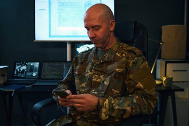 Kamuflaj üniformalı asker ofis ortamında görev başında, elinde bir şeye odaklanıyor, arka planda monitörler ve evrak işleriyle profesyonel bir ortam yaratıyor.