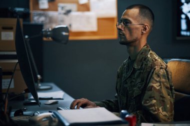 Askeri personel iş yerindeki veri analiziyle meşgul, kamuflaj üniforması giyiyor ve bilgisayar ekranına odaklanmış durumda.