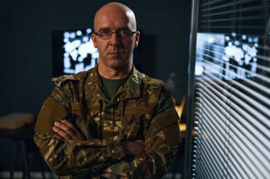 Kamuflaj üniformalı bir askerin portresi komuta merkezinde ekran ve ekipmanlarla dolu bir şekilde duruyor. Kararlılık ve odaklanma gösteren ciddi ifade