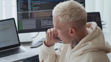 Ofis masasında uygulama güncellemesi üzerinde çalışırken cep telefonuyla konuşan dövmeli genç erkek programcının yan görüntüsü