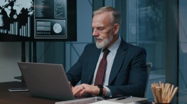 Orta boy, beyaz takım elbiseli bir işadamının, finans departmanındaki masada oturan kablosuz bir dizüstü bilgisayarla konuşurken görüntüsü.