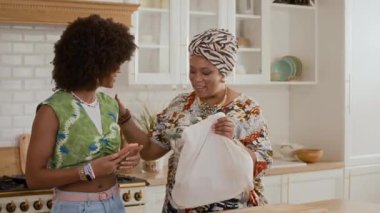 Orta boy Afro-Amerikalı genç bir kızın akıllı telefon kullanarak annenin mutfak çantalarını boşaltmasına yardım ettiği bir fotoğraf.