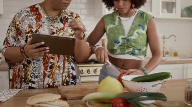 Ev mutfağında birlikte yemek pişirirken iki kişilik çok ırklı ailenin dijital tablette tarifini kullanmasının el kamerası görüntüsü.