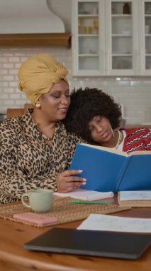 Çok ırklı anne ve kızının evde mutfaktaki masada otururken kitap okuduğu dikey çekim.