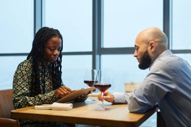Restoranda panoramik pencereli bir şekilde otururken menüyü inceleyen çok ırklı bir çift.