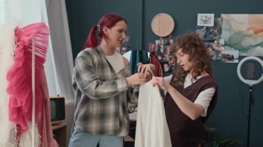 Kızıl saçlı kız, beyaz bluz ve pantolon giyerken kız arkadaşının kıyafet seçmesine yardım ediyor.