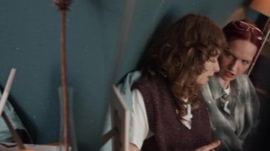 Dijital tablet ve akıllı telefon taşıyan iki beyaz kadın duvara yaslanırken birbirleriyle konuşuyorlar.