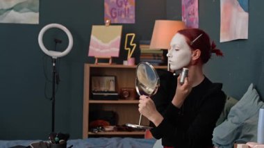 Kızıl saçlı Z geninin yatağının yanındaki masadan makyaj aynasını alırken ve yatak odasında otururken kulak ve boynuna beyaz bir temel atarken belden yukarı çekilen fotoğrafı.